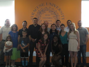 16 persons are posed in front of a wall with the Syracuse University trademark on it