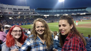 Three women pose seated in a baseball stadium during a game