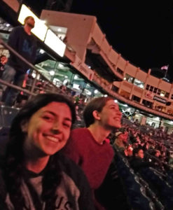 2 women are smiling and seated in a baseball stadium during a game at night